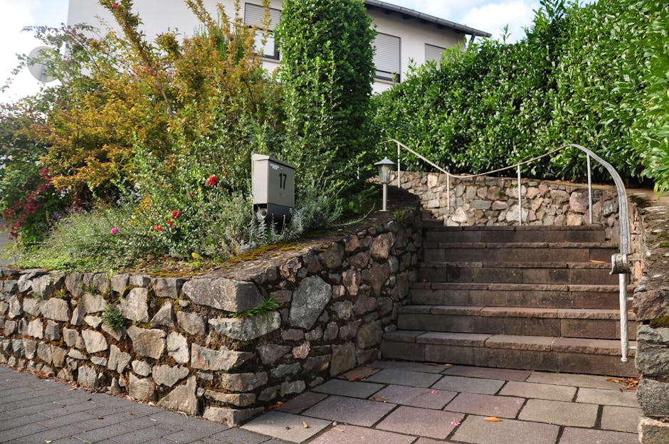 A narrow, uneven garden pathway with a mixture of dirt and fallen leaves, bordered by a green wooden fence on the left and a metal handrail on the right. The path is surrounded by tall, leafless trees