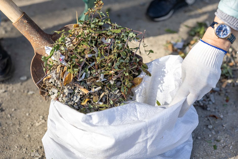 A close-up view of a person wearing a light-colored glove and a wristwatch, using a shovel to place a mixture of garden debris, including small twigs, leaves, and bits of concrete or gravel, into a la