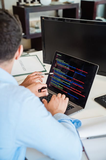 A man with glasses and a beard is seated at a wooden desk, working on a dual-monitor computer setup. The left monitor displays a dark-themed file management interface with folders and filenames, while